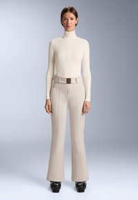 Priliehavý smotanový top s dlhým rukávom a polozipsom, kombinovaný s béžovými zvonovými nohavicami s detailami na švoch a zodpovedajúcim opaskom. Čierne textúrované čižmy.