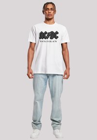 White cotton t-shirt featuring a black AC/DC "Back In Black" logo. Paired with light blue denim jeans and white sneakers.