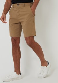 Shorts en coton beige avec une taille plate, des poches latérales et une fermeture à bouton, associés à des baskets blanches avec des accents noirs.