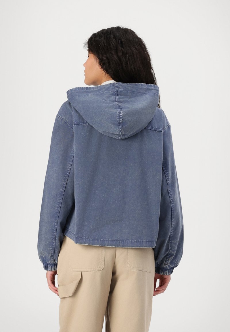 Veste en denim bleu délavé avec capuche, poignets élastiques et design court. La texture semble douce et légèrement usée.