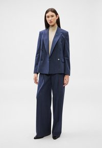 Žena stojící, oblečená v tmavě modrém dvouřadém saku, odpovídajících širokých kalhotách, béžovém roláku a černých špičatých botách, na jednobarevném pozadí.