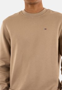 Sudadera beige hecha de una tela texturizada, con un cuello redondo y un pequeño logo bordado en el lado izquierdo del pecho.