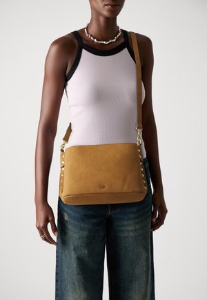 Femme portant un débardeur blanc avec bordure noire, un jean bleu, un collier de perles et un sac bandoulière beige orné de clous dorés.