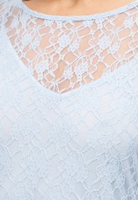 Κοντινό πλάνο γαλάζιου δαντελένιου υφάσματος με λουλουδένια σχέδια στο ντεκολτέ και την περιοχή του στήθους ενός ένδυματος.