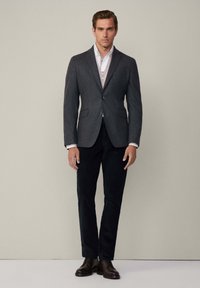Blazer grigio scuro su misura con revers a punta, abbinato a una camicia bianca con colletto. Il modello indossa pantaloni scuri e scarpe in pelle marrone.