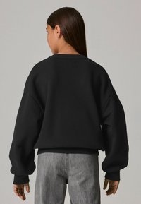 Sudadera negra de gran tamaño con hombros caídos, cuello y puños acanalados, combinada con jeans gris claro. Textura de tela suave.