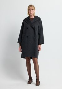 Mujer vestida con un abrigo gris oscuro de doble botonadura y gran cuello, medias negras transparentes y zapatos T-strap marrones, de pie contra un fondo liso.