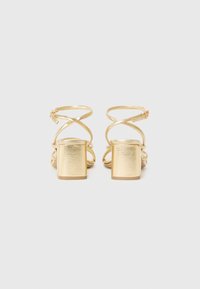 Gold heeled sandals with cross-strap design, textured metallic finish, block heel, and adjustable ankle straps.