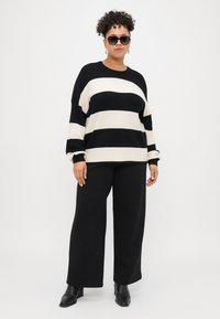 Svart og kremstripet strikkegenser med vide ermer, kombinert med svarte vide bukser og svarte ankelstøvler; fullført med solbriller.