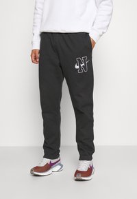Svarta sweatpants med vitt "N"-logotyp, avslappnad passform, elastisk midja, kombinerad med mångfärgade löparskor med lila accenter.
