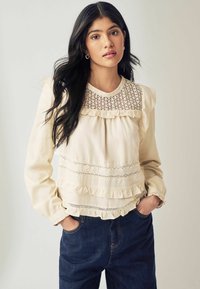 Cremefarvet bluse med blonder, flæser og lange ærmer. Sammen med mørk denim jeans. Glat tekstur.