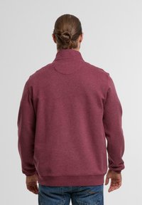 Burgundi színű fleece pulóver magas gallérral, bordázott mandzsettákkal és aljjal. Sima tapintás, laza szabás és minimalista varrási részletek.