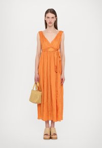 Orange, krinklad maxiklänning med V-ringning och midjeband. Modellen håller en vävd handväska i beige och bär platåskor.