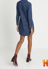 Denim-mekko, jossa on pitkät hihat, kaareva helma, etupaneelin yksityiskohta ja polven puoliväliin ulottuva pituus. Yhdistetty kiiltäviin burgundinpunaisiin nilkkabootseihin.