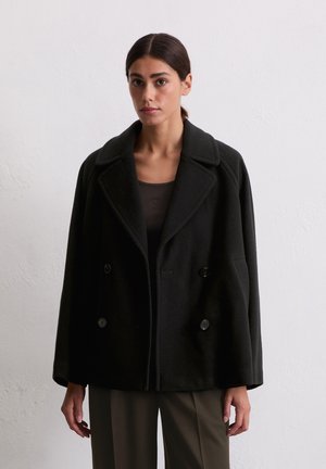 Mujer con cabello oscuro en una coleta baja, que lleva un abrigo negro de doble botonadura suelto sobre una blusa oscura y pantalones marrones de pierna ancha, frente a una pared blanca.