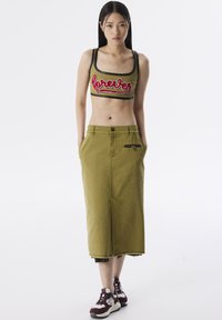 Jupe en denim vert olive avec fente avant et poches lat�érales, associée à un crop top ajusté vert olive ornée d'une broderie rouge.