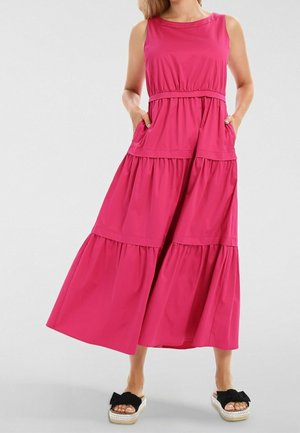 Woman in sleeveless pink tiered maxi dress with pockets, wearing black bow slip-on platform sandals, standing against plain background.