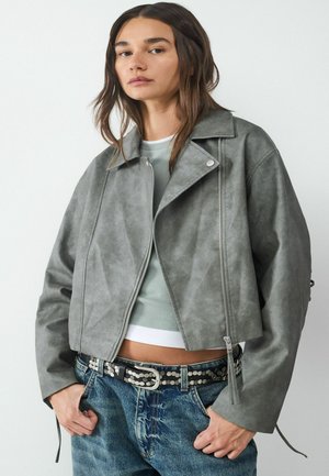 Kragen van grijs nepleer jack met brede kraag, voorzien van ritssluiting en drukknoopsluiting. Model draagt een lichtblauw shirt en denim jeans met een gesp met studs.