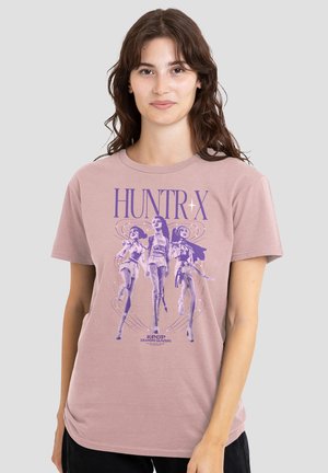 Frau trägt ein mauvefarbenes T-Shirt mit lila Grafik von drei weiblichen K-Pop-Dämonenjägern und dem Text „HUNTR X“ und „K-POP DEMON HUNTERS.“