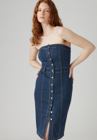 Vestido de mezclilla sin tirantes en azul oscuro, con botones frontales, una silueta ajustada y un corte a la altura de la rodilla con una abertura lateral.