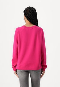 Fuksjowa bluza o luźnym kroju, z raglanowymi rękawami i ściągaczami u rękawów. Materiał wydaje się miękki, o gładkiej fakturze i bez widocznych wzorów.