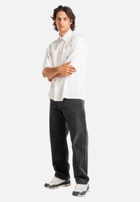 Weißes kurzärmliges Button-up-Hemd, schwarze Cordhose und graue Sneakers. Modell steht mit verschränkten Armen und blickt nach vorne.
