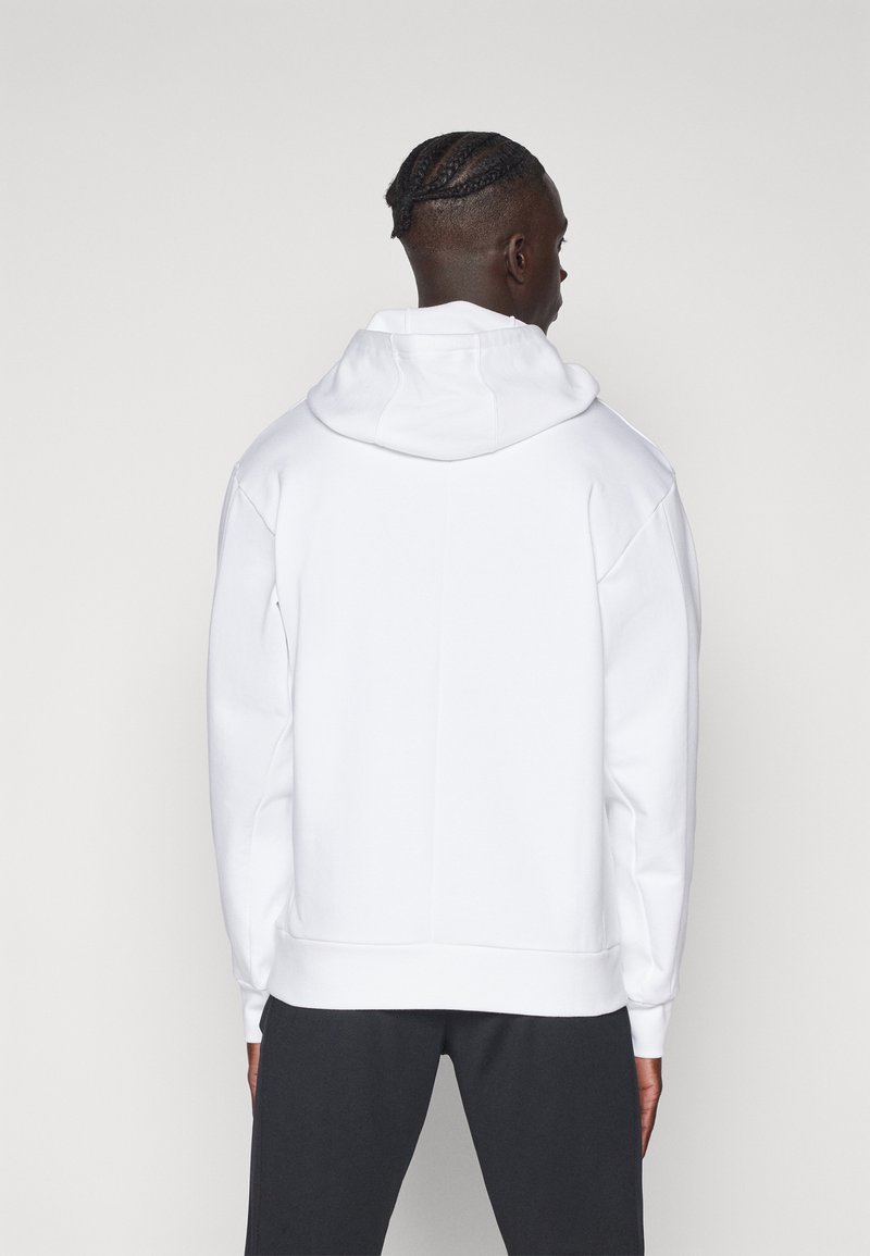 Sudadera blanca con capucha y hombros caídos. Presenta un dobladillo y puños acanalados, con una textura suave y gruesa de algodón. Diseño simple, sin patrones.