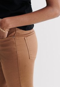Pantalones de algodón marrón con un diseño ajustado. Cuentan con un bolsillo lateral y detalles de costura a lo largo de la cinturilla y los bordes del bolsillo.