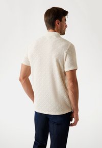 Polo majica kratkih rukava u krem boji s teksturiranim dijamant uzorkom. Ima klasičan ovratnik i ravni rub. Nositi uz tamne traperice.
