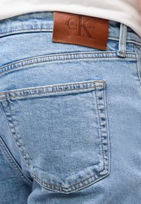 Τζιν τζην ανοιχτού μπλε χρώματος πίσω τσέπη με καφέ δερμάτινο λογότυπο Calvin Klein κοντά στη μέση κάτω από λευκό ύφασμα.