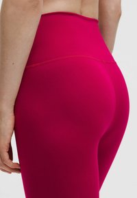 Záber zblízka na osobu nosiacu legíny z vysoko pásového jasne ružového materiálu, zobrazujúci podrobnosti prešitého švu na zadnej a bočnej strane.