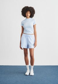 Σετ αθλητικών ρούχων σε ανοιχτό μπλε χρώμα, που περιλαμβάνει εφαρμοστό κοντομάνικο τοπ και φαρδιά σορτς με κορδόνι στη μέση. Το υλικό φαίνεται μαλακό και διαθέτει λεπτομέρεια με λογότυπο.