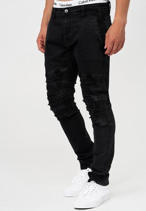 Mężczyzna noszący czarne podarte obcisłe dżinsy, widoczny biały pas marki Calvin Klein oraz białe trampki, stojący na jednolitym tle.