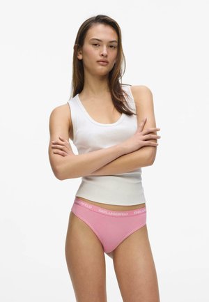 Vrouw staand met gekruiste armen, draagt een wit tanktop en roze Karl Lagerfeld ondergoed tegen een witte achtergrond.