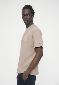 Καφετί κοντομάνικη μπλούζα με στρογγυλή λαιμόκοψη, με γραφικό σχέδιο στο μπροστινό μέρος και μικρό λογότυπο στο μανίκι. Μαλακό βαμβακερό ύφασμα.