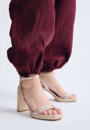 Pies con sandalias de tacón beige con correas en el tobillo combinadas con pantalones sueltos burdeos con puños elásticos.