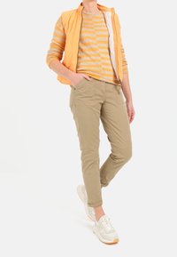 Leichtes oranges Weste über einem gestreiften beige-gelben Oberteil, khakifarbene Hose mit Taschen und cremfarbene Sneaker mit strukturierten Akzenten.