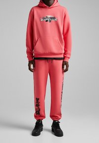 Korallrosa huvtröja med grafisk design, kenguruficka och matchande mjukisbyxor med svarta detaljer och text. Avslutas med svarta skor.