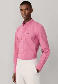 Mann trägt ein pinkes, langärmliges Hemd mit Knöpfen und Brusttasche sowie weiße Hose, steht mit einer Hand in der Tasche vor hellem Hintergrund.