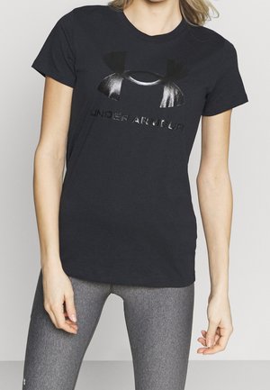 Kvinde iført en sort Under Armour t-shirt og grå leggings, stående med armene afslappet ved siden mod en ensfarvet baggrund.