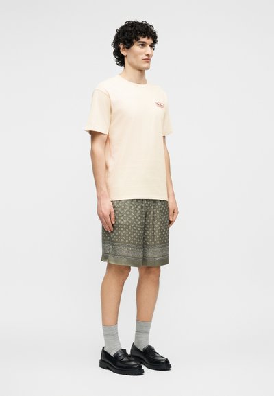 Νεαρός άνδρας με σγουρά μαλλιά που φοράει μπεζ κοντομάνικο T-shirt, πράσινα σορτς με σχέδια, γκρι κάλτσες και μαύρα μοκασίνια, στέκεται πλαγίως.