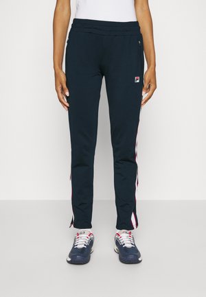 Person trägt marineblaue Sporthose mit weißen und roten Seitenstreifen, Logo auf dem linken Oberschenkel und marineblaue und weiße Turnschuhe.