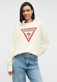 Γυναίκα με κρεμ πουλόβερ που φέρει το κόκκινο και μαύρο τρίγωνο λογότυπο της Guess και τζιν ψηλής μέσης ανοιχτού μπλε χρώματος με ζώνη, μπροστά σε απλό φόντο.