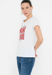 T-shirt bianco in cotone con un design grafico colorato rosso sulla parte frontale. Maniche corte, vestibilità comoda e scollatura rotonda.