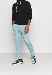 Felpa nera con logo bianco, pantaloni jogger azzurri chiari e scarpe sportive in tonalità neutre con accenti neri e dorati.