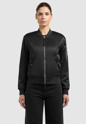 Femeie purtând o jachetă bomber din satin negru cu fermoar frontal, buzunare laterale și un buzunar cu fermoar pe mâneca stângă, asortată cu pantaloni negri.