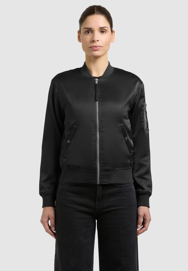 Femme portant une veste bomber en satin noire avec fermeture éclair frontale, poches latérales et une poche zippée sur la manche gauche, associée à un pantalon noir.
