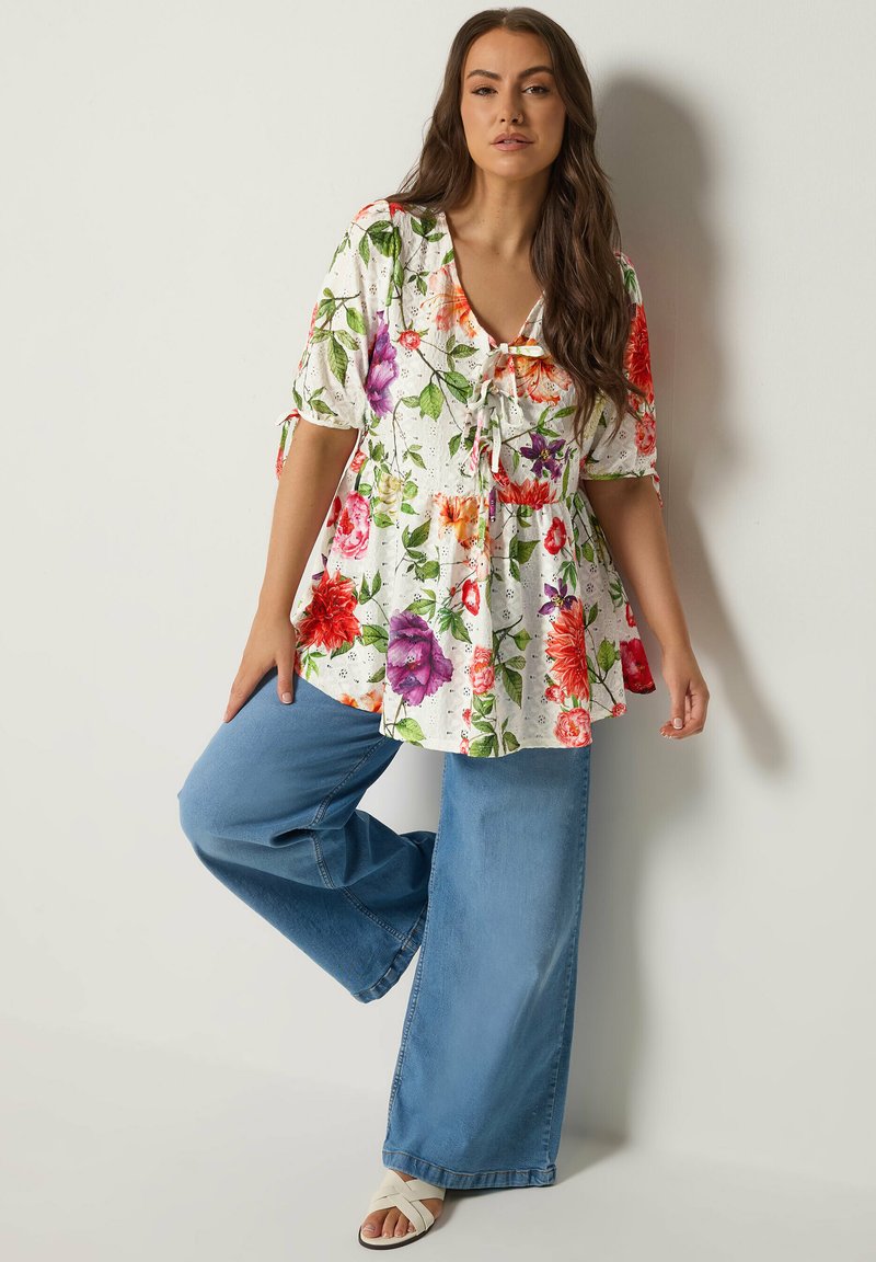 Frau trägt eine weiße Blumenbluse mit Bindedetails und weit geschnittene blaue Jeans, posiert mit einem angewinkelten Bein vor einem hellen Hintergrund.
