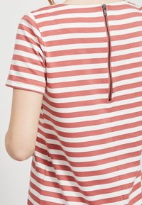 Pasiasta koszulka w różowym i białym kolorze, wykonana z bawełnianego materiału, z krótkimi rękawami i zamkiem błyskawicznym z tyłu. Gładka faktura i luźny krój.