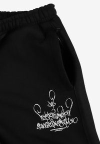 Czarne spodnie dresowe z elastycznym paskiem na sznurki, bocznymi kieszeniami na zamek błyskawiczny i srebrnym logo w stylu graffiti na udzie. Miękka faktura materiału.